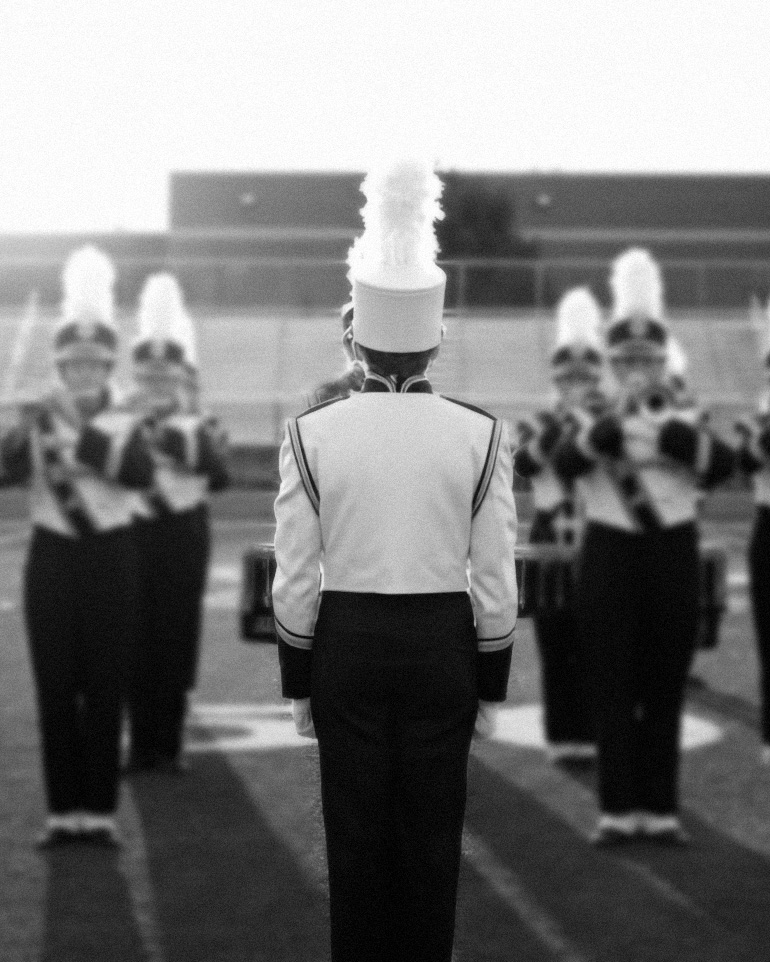 A drum major stands with their back to the camera, facing a high school marching band held at attention in formation on a football field.